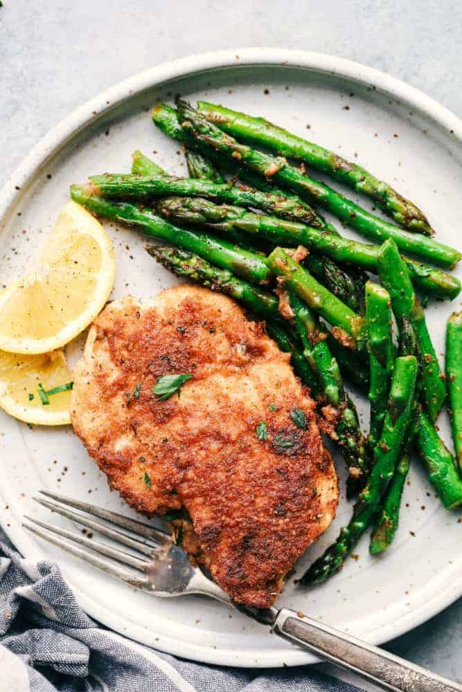 Crispy Garlic Chicken with Asparagus is an easy 30 minute meal with crispy breaded chicken and crisp and tender asparagus. This is a family favorite meal! One piece of crispy garlic chicken with asparagus and sliced lemon on a white plate.