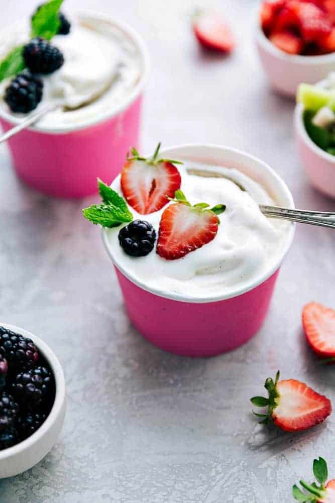 This vanilla soft serve frozen yogurt is delicious and perfect for the hot summer days! Everyone goes crazy for it! Two pink paper cups of vanilla soft serve frozen yogurt. One of them has fresh strawberries on top and the other has blackberries on top.