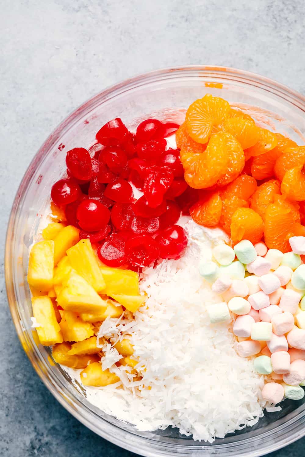 Ambrosia Salad is filled with mandarin oranges, pineapple, maraschino cherries, coconut, and marshmallows and mixed in a fluffy cool whip! This is a potluck classic! Ambrosia Salad ingredients in a glass bowl.