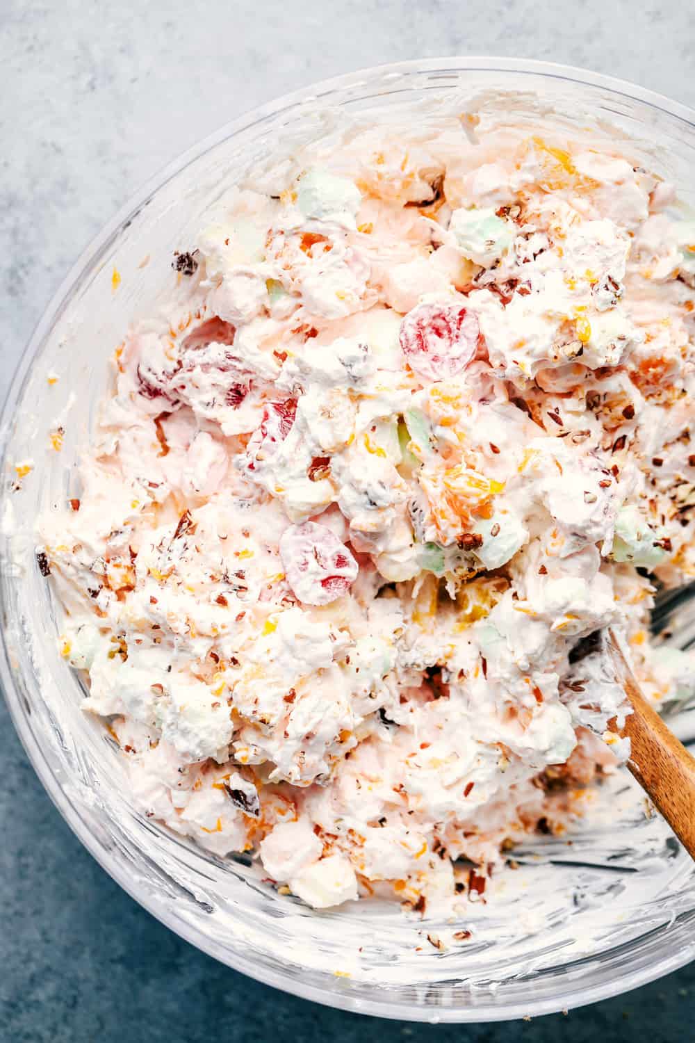 Ambrosia Salad is filled with mandarin oranges, pineapple, maraschino cherries, coconut, and marshmallows and mixed in a fluffy cool whip! This is a potluck classic! Ingredients being mixed in a glass bowl with a wooden spoon.