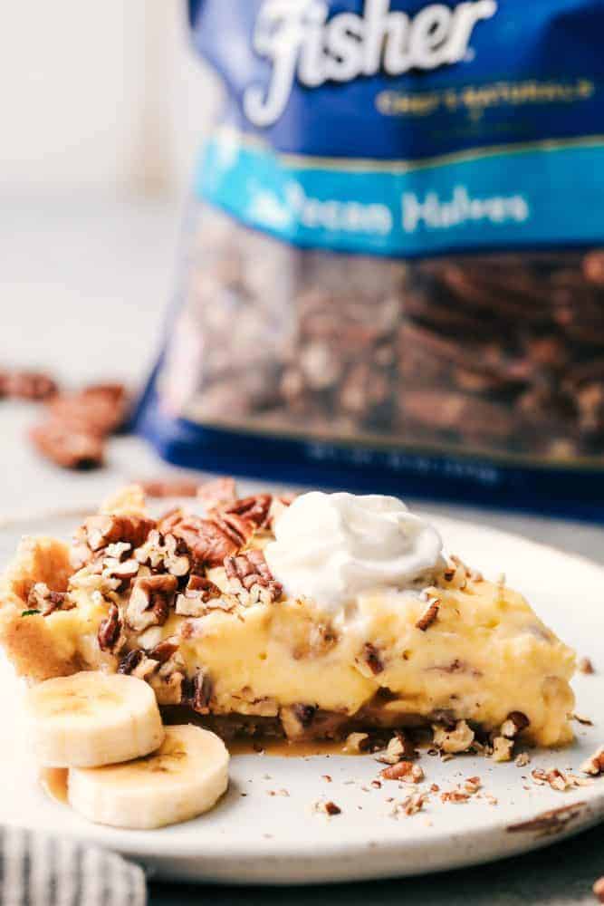 No Bake Banana Caramel Pecan Pie is made with a shortbread crust topped with caramel and a creamy banana filling with crunchy pecans. This makes one incredible no bake pie! A slice of No Bake Banana Caramel Pecan Pie on a white plate with a couple of slices of fresh cut banana. There is a back of Fisher pecan halves in the background.