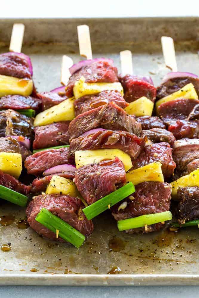 Teriyaki Beef Skewers are an easy to make grilled appetizer or entree. Tender pieces of sirloin steak marinated in an Asian inspired sauce and cooked on a hot grill until lightly charred. Each beef skewer is drizzled with a sweet and savory teriyaki sauce and garnished with sesame seeds, green onion, and cilantro. Uncooked Teriyaki Beef Skewers on a cooking sheet.