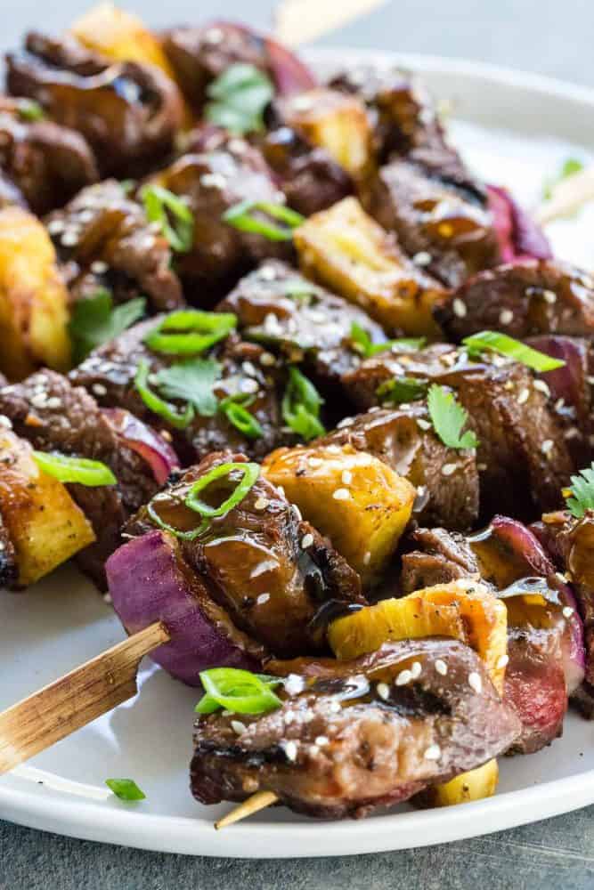 Teriyaki Beef Skewers are an easy to make grilled appetizer or entree. Tender pieces of sirloin steak marinated in an Asian inspired sauce and cooked on a hot grill until lightly charred. Each beef skewer is drizzled with a sweet and savory teriyaki sauce and garnished with sesame seeds, green onion, and cilantro. Teriyaki Beef Skewers on a white plate.