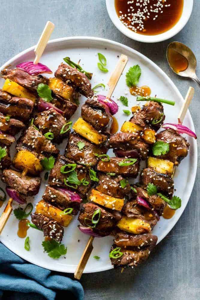 Teriyaki Beef Skewers are an easy to make grilled appetizer or entree. Tender pieces of sirloin steak marinated in an Asian inspired sauce and cooked on a hot grill until lightly charred. Each beef skewer is drizzled with a sweet and savory teriyaki sauce and garnished with sesame seeds, green onion, and cilantro. Teriyaki Beef Skewers on a white plate with extra teriyaki sauce in a small dish.