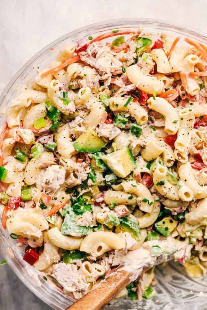 Tuna Avocado Macaroni Salad is an AMAZING creamy macaroni salad that is filled with chopped veggies, avocado, and tuna. This is going to impress everyone at your next potluck! Close up of Tuna Avocado Macaroni Salad in a clear bowl with a wooden spoon for serving.