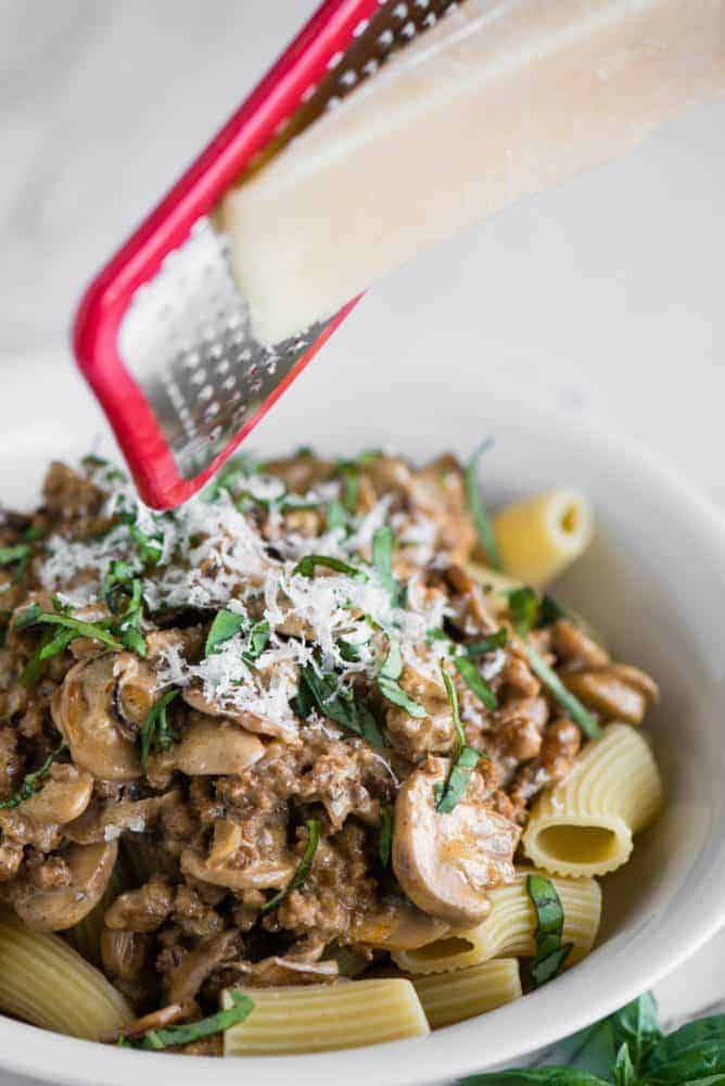 Creamy Sausage Mushroom Rigatoni is a simple Italian pasta recipe that makes for an easy and delicious dinner in less than 30 minutes! This meal isย is pure comfort food that the entire family will love. Cheese being freshly grated on top of Creamy Sausage Mushroom Rigatoni in a white bowl.