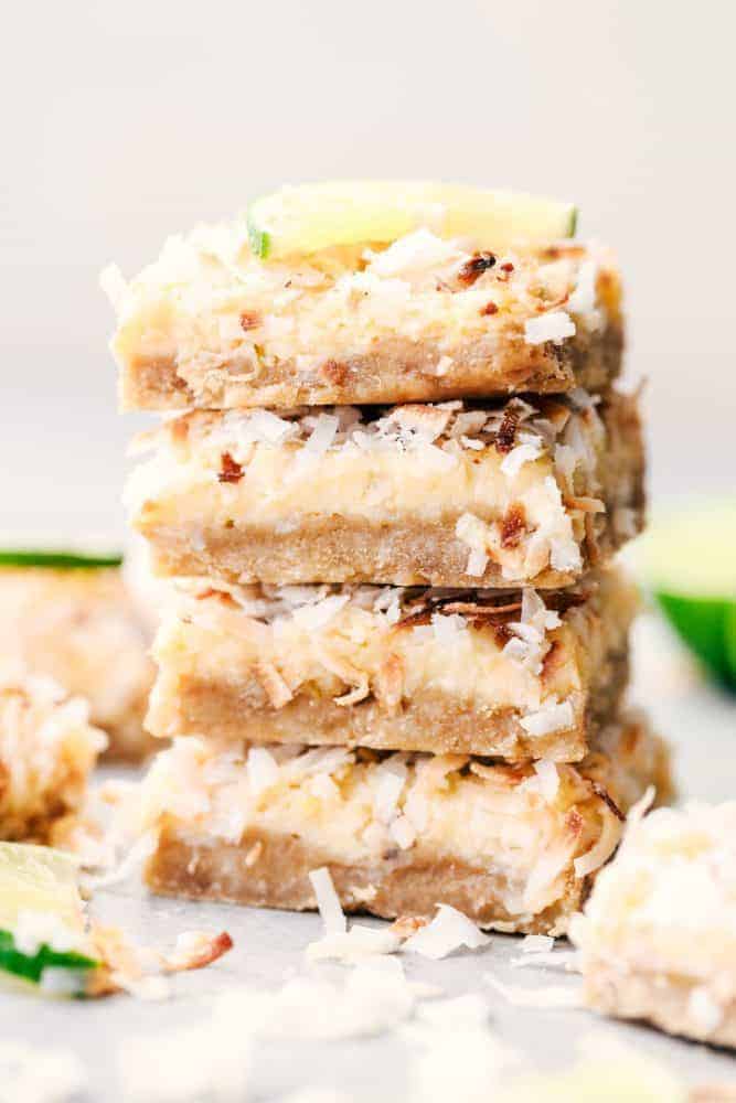 Coconut Lime Butter Bars have an amazing buttery brown sugar crust with a lime filling and topped with coconut flakes. These are the perfect cool and creamy summer bars!ย Four Coconut Lime Butter Bars stacked on top of each other with a fresh sliced lime on top.