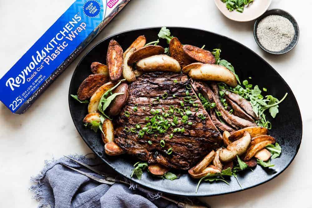 Brown Sugar Garlic Flank Steak is made with the most amazing brown sugar garlic soy marinade. The steak is grilled to perfection and melt in your mouth delicious!ย Brown Sugar Garlic Flank Steak on a black serving platter with a box of Reynolds Plastic Wrap next to it.