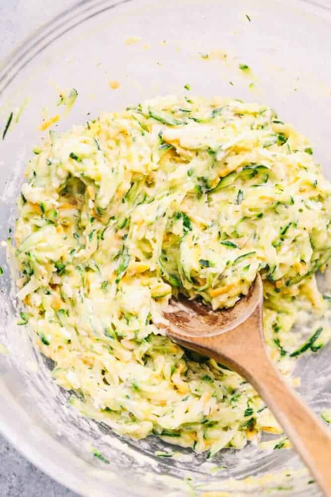 Cheesy Zucchini Fritters are filled with three cheeses and zucchini. These are savory and easy to make and the perfect way to use up that garden zucchini! Mix for Cheesy Zucchini Fritters in a clear bowl with a wooden spoon.