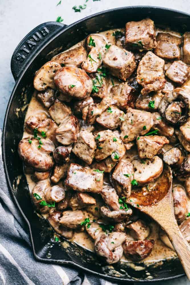Creamy Garlic Steak Bites with Mushrooms are made in the most amazing creamy garlic sauce with melt in your mouth steak bites and mushrooms! Creamy garlic streak bites with mushrooms cooking in a staub pan with a wooden spoon stirring it.