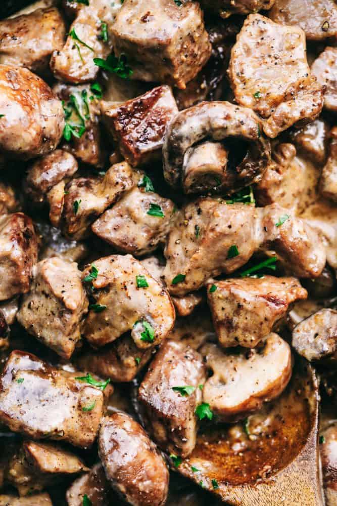 Creamy Garlic Steak Bites with Mushrooms are made in the most amazing creamy garlic sauce with melt in your mouth steak bites and mushrooms! Up close photo of garlic steak bites with mushrooms and a wooden spoon in the lower right hand corner.