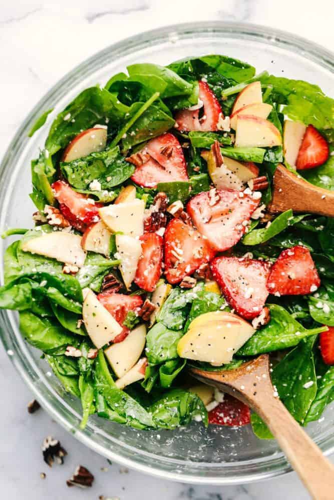 Strawberry, Apple, and Pear Spinach Salad is an easy to make spinach salad with crisp apples, pears and strawberries hidden throughout. Topped with feta cheese, crunchy pecans and tossed in a delicious apple cider dressing! Strawberry, Apple, and Pear Spinach Salad in a clear bowl with a wooden spoon.