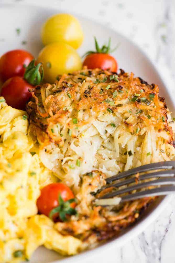 Baked Parmesan Hashbrowns, baked to golden brown perfection and made with freshly grated potatoes, are a breakfast favorite! They are loaded with flavor and are the perfect addition to your hearty breakfast. If you love traditional hashbrowns, then you will really love these Baked Parmesan Hashbrowns! Baked Parmesan Hashbrowns on a white plate with scrambled eggs and small tomatoes.