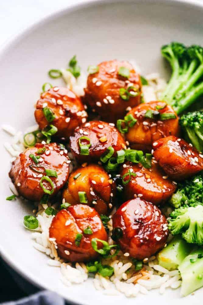 Honey Garlic Butter Scallops get pan seared and then coated in the most amazing honey garlic butter sauce. This meal is so quick and easy that you will make it again and again!ย Honey Garlic Butter Scallops over white rice in a white bowl with broccoli on the side.