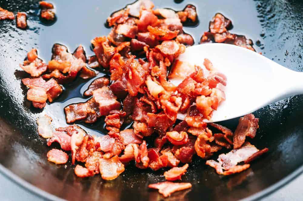 Bacon Fried Rice is 1000x better than takeout and has crispy bacon, eggs, peas, carrots and rice that combine to make the best fried rice ever! Bacon being fried in a frying pan using a wooden spoon.