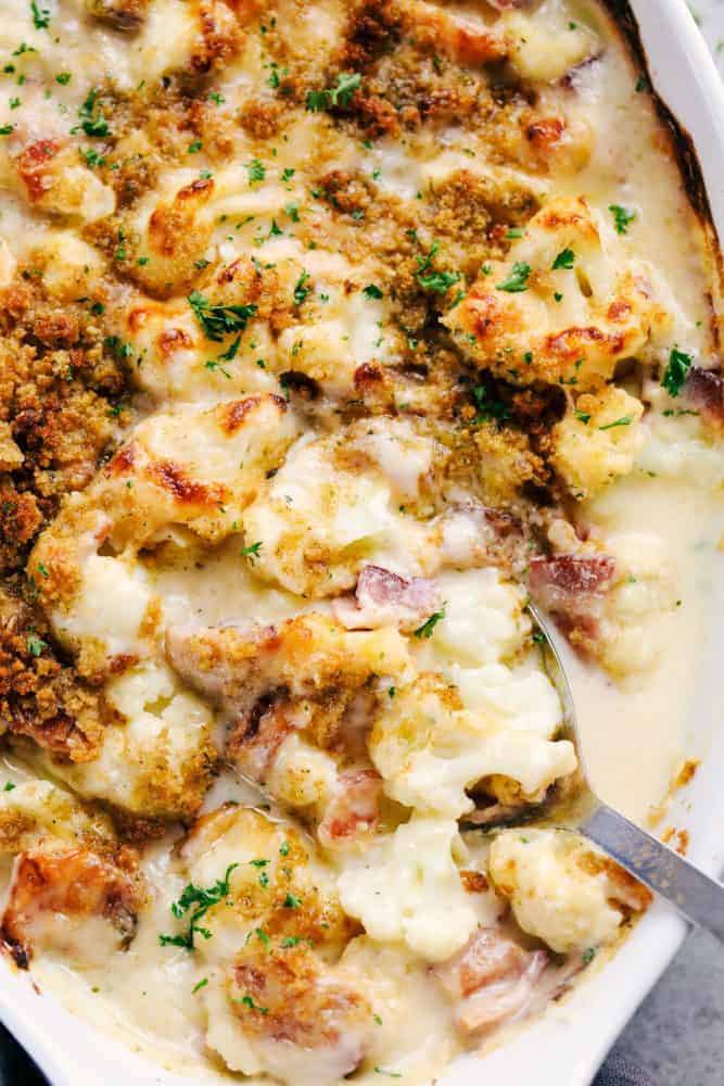 Creamy Cauliflower Au Gratin with Bacon makes such an amazing side dish. It is filled with tender cauliflower and bacon in a cheesy sauce that is topped with bread crumbs. This will be the hit of the dinner table! Creamy cauliflower au gratin with bacon in a white casserole dish with a silver spoon pulling up part of the cauliflower and au gratin with bacon to show the chunks.