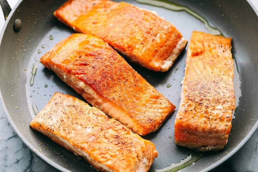 Creamy Tuscan Garlic Salmon is an easy 30 minute restaurant quality meal. The pan seared salmon gets smothered in the most amazing creamy tuscan garlic sauce with sun dried tomatoes and spinach. This is one unforgettable meal! Salmon fillets being cooked in a frying pan getting ready for the creamy Tuscan garlic sauce.