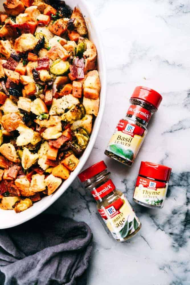 Roasted Autumn Vegetable Stuffing is the perfect side dish for your holiday meal. Filled with roasted autumn vegetables and bacon, this is the perfect addition to the dinner table. Roasted autumn vegetable stuffing and a white casserole dish with McCormick seasoning's on the side are the bottles of basil leaves, sage and thyme and their bottles.