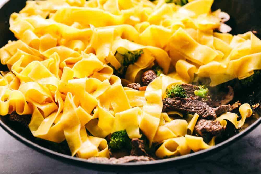 Garlic Beef and Broccoli Noodles is made with tender melt in your mouth beef in the most amazing garlic sauce. Add some mushrooms, broccoli and noodles for an amazing meal in one! Thick egg noodles are being placed in the skillet with garlic beef and broccoli.