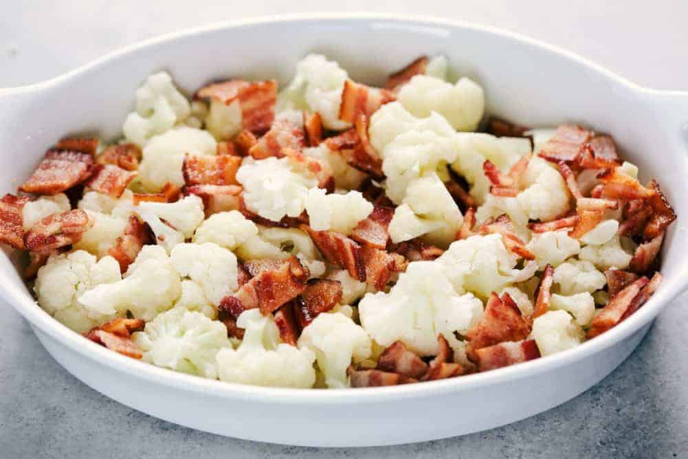 Creamy Cauliflower Au Gratin with Bacon makes such an amazing side dish. It is filled with tender cauliflower and bacon in a cheesy sauce that is topped with bread crumbs. This will be the hit of the dinner table! Chopped up cauliflower with Smithfield bacon pieces cut up in a white casserole dish being ready to bake.