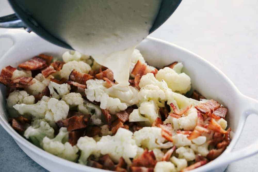 Creamy Cauliflower Au Gratin with Bacon makes such an amazing side dish. It is filled with tender cauliflower and bacon in a cheesy sauce that is topped with bread crumbs. This will be the hit of the dinner table! Chopped up a cauliflower and Smithfield bacon pieces in a white casserole dish having the au gratin being poured over top is it from the sauce pan.