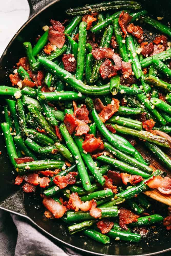 Garlic Parmesan Green Beans with Bacon is such a delicious and classic side dish. Tender green beans cooked in garlic and parmesan with added bacon will be a winner at the dinner table! Garlic Parmesan green beans with bacon and a black skillet with the wooden spoon stirring.