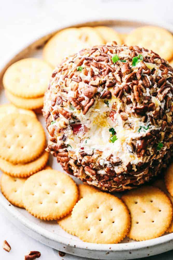 Pineapple Cheese Ball is the next best twist on a favorite classic. With Different textures and flavors it will be the hit at your next gathering! Pineapple cheese bar with a bite out of the top of it to show the pineapple and chives With Ritz crackers surrounding it served on a white plate.