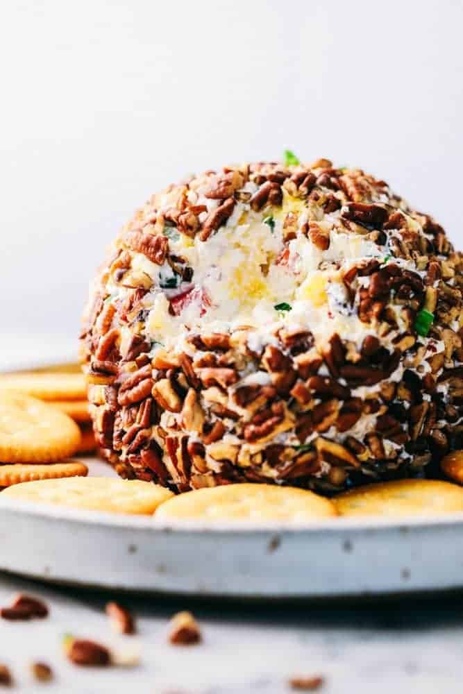 Pineapple Cheese Ball is the next best twist on a favorite classic. With Different textures and flavors it will be the hit at your next gathering! Pineapple cheese ball with crackers all around it on a white plate.