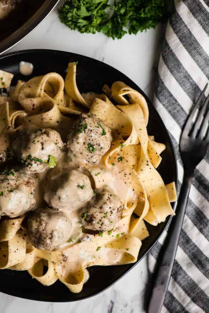 Swedish Meatball Pasta combines flavorful homemade pork and beef Swedish meatballs with a heavenly cream sauce and perfectly cooked pasta. Swedish Meatball pasta on a black plate with a black fork on a napkin next to the plate on the righthand side.