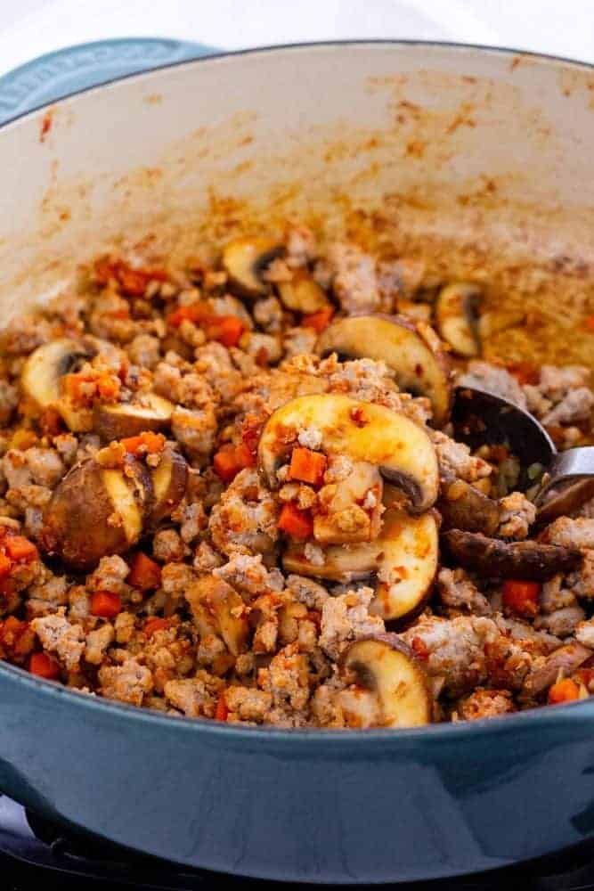 Turkey Bolognese with Roasted Spaghetti Squash is a hearty Italian feast with a low carb twist. Ground turkey is gently simmered with carrots, onions, and mushrooms in a rich tomato sauce. The flesh of the squash separates into strands to mimic pasta noodles for a tasty vegetable-packed meal. ground turkey, diced carrots and mushrooms cooking in a dutch oven