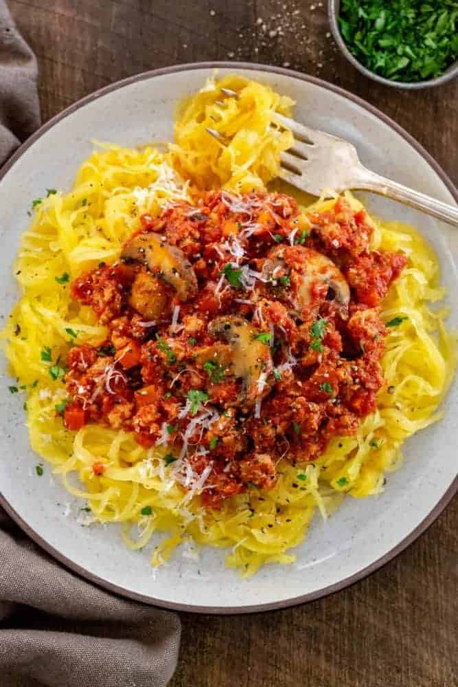 Turkey Bolognese with Roasted Spaghetti Squash is a hearty Italian feast with a low carb twist. Ground turkey is gently simmered with carrots, onions, and mushrooms in a rich tomato sauce. The flesh of the squash separates into strands to mimic pasta noodles for a tasty vegetable-packed meal. top down photo of a plate of turkey meat sauce over a bed of spaghetti squash vegetable noodles