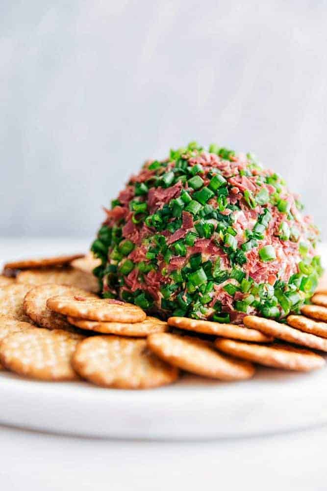 Chipped Beef Cheese Ball is such a classic and amazing cheeseball filled with cheddar cheese, cream cheese, green onions, and dried beef. This easy cheese ball is going to be the hit at any gathering!ย Chipped beef cheese ball on a white plate with crackers surrounding it.