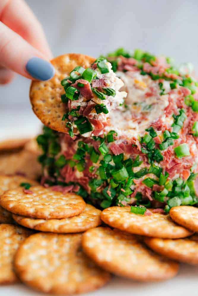 Chipped Beef Cheese Ball is such a classic and amazing cheeseball filled with cheddar cheese, cream cheese, green onions, and dried beef. This easy cheese ball is going to be the hit at any gathering!ย Chipped beef cheese ball with a hand dipping a cracker into it.