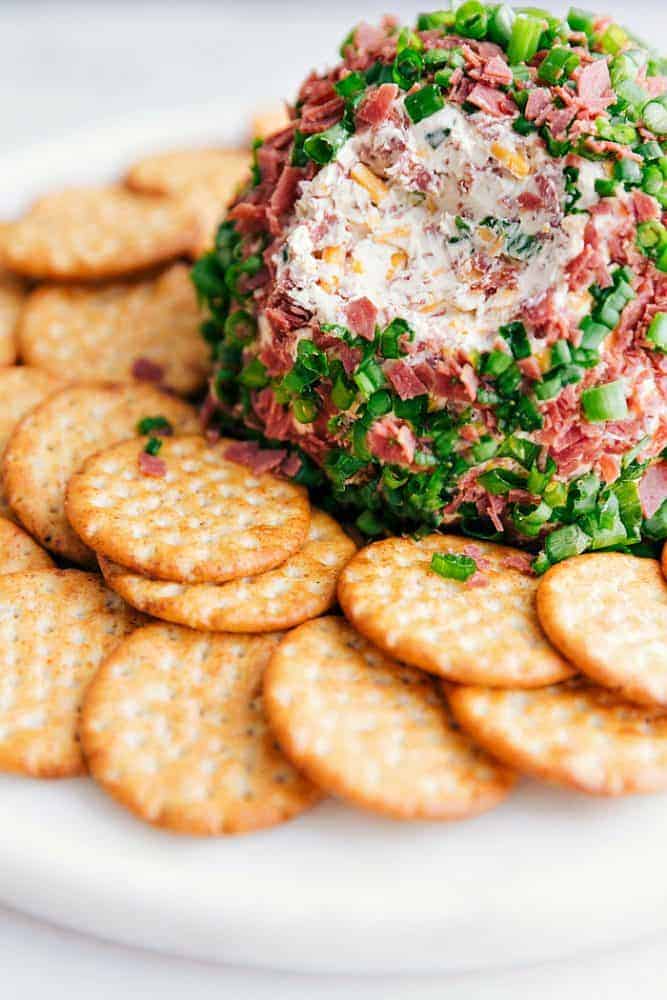 Chipped Beef Cheese Ball is such a classic and amazing cheeseball filled with cheddar cheese, cream cheese, green onions, and dried beef. This easy cheese ball is going to be the hit at any gathering!ย Chipped beef cheese ball on a white plate with crackers surrounding it.