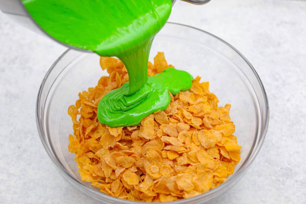 Christmas Cornflake wreaths are a fun, whimsical treat that symbolizes infinite love during the holiday season. Crunchy cornflakes, bright green food coloring, and a sweet, gooey glaze combine to make the cutest Christmas wreath treats. Angle shot of someone pouring the green marshmallow mixture into the cornflakes in a glass bowl.