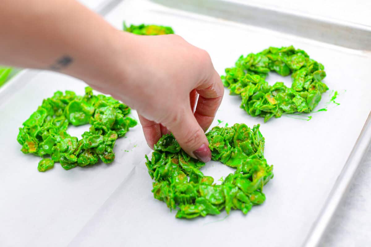 Christmas Cornflake wreaths are a fun, whimsical treat that symbolizes infinite love during the holiday season. Crunchy cornflakes, bright green food coloring, and a sweet, gooey glaze combine to make the cutest Christmas wreath treats. Angle shot of someone shaping the wreaths with their hands on parchment paper on a cookie sheet.