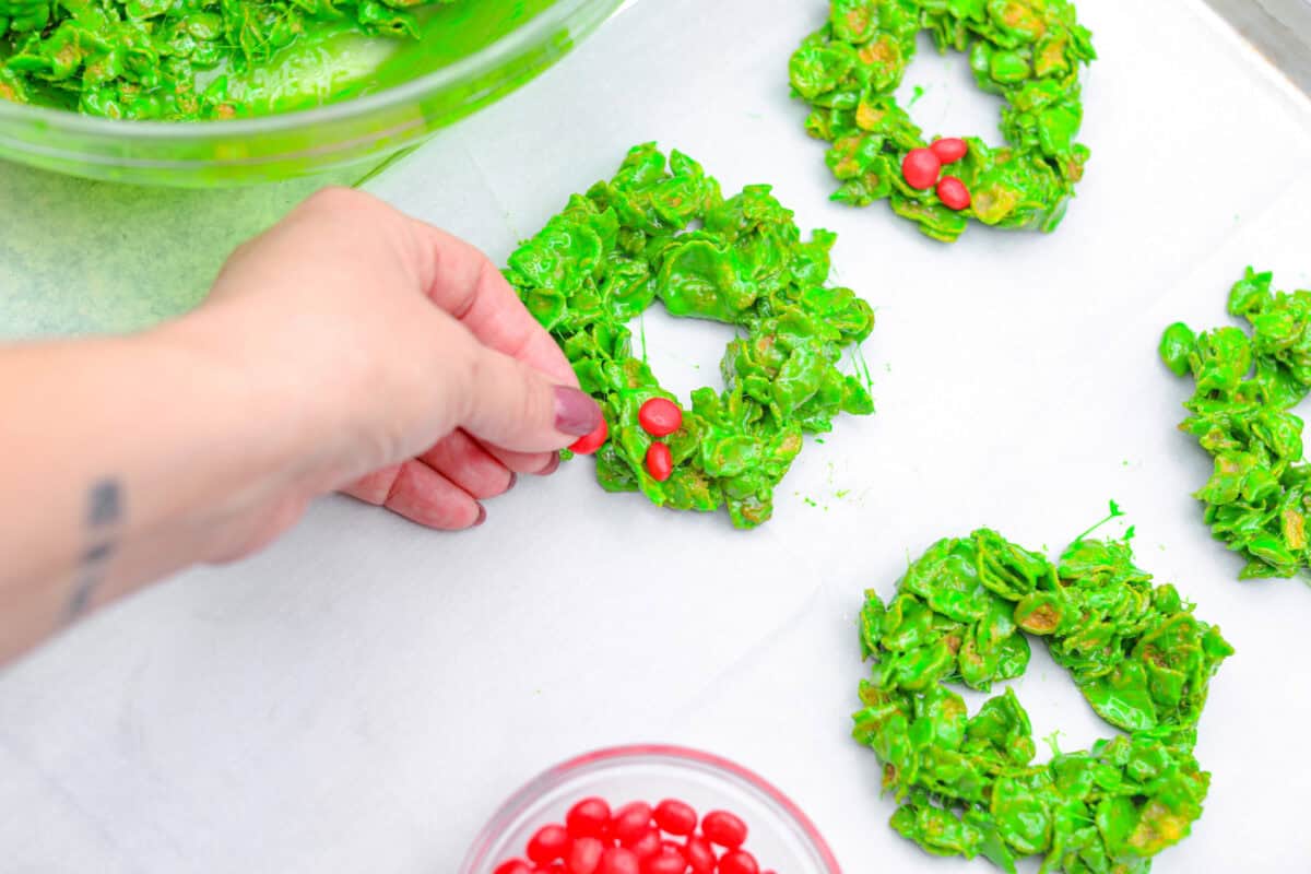 Christmas Cornflake wreaths are a fun, whimsical treat that symbolizes infinite love during the holiday season. Crunchy cornflakes, bright green food coloring, and a sweet, gooey glaze combine to make the cutest Christmas wreath treats. Overhead shot of someone placing the cinnamon candy on the wreaths.