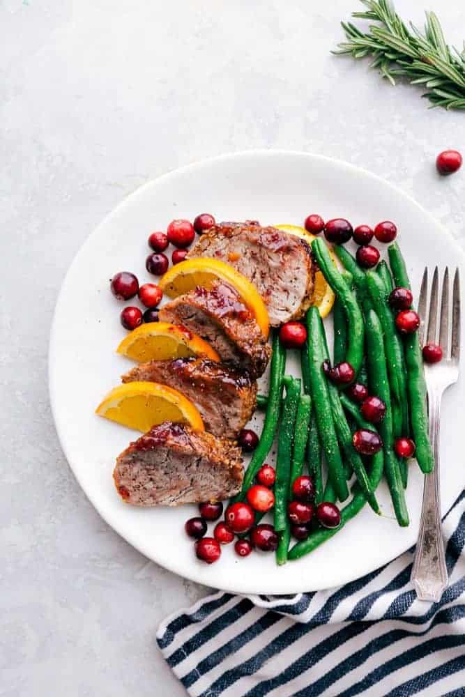 Roasted Cranberry Orange Pork Tenderloin with Green Beans is such a flavorful and easy meal. The cranberry orange glaze is perfect on top of the pork and green beans. This will be the star of your holiday dinner table! Roasted cranberry orange pork tenderloin with green beans on a white plate with a metal fork and a black and white cloth on the side.