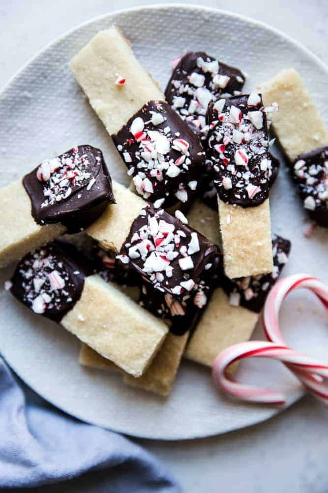 Chocolate Peppermint Dipped Shortbread Cookies are buttery, melt in your mouth shortbread cookies that get dipped in chocolate and sprinkled with crushed peppermint. These cookies make the perfect holiday treat! Close up photo of chocolate peppermint dip shortbread cookies on a white plate with two candy canes on the side.