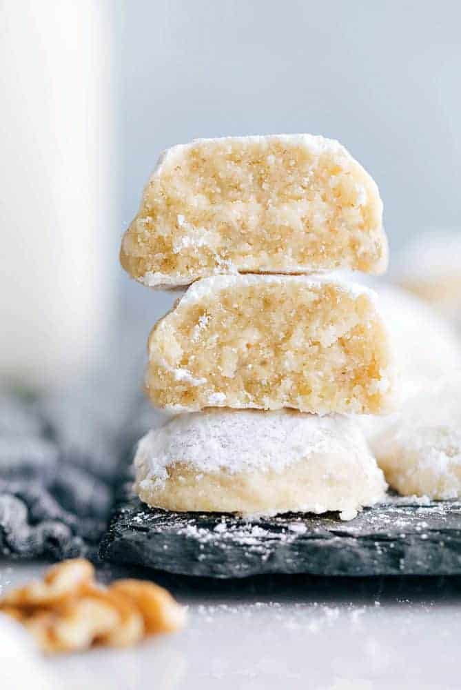 Snow Kiss Cookies are buttery cookies with walnuts that just melt in your mouth. They get dusted in powdered sugar and are are such an amazing classic cookie! Snow kiss cookies stacked on top of one another on a black cutting board.
