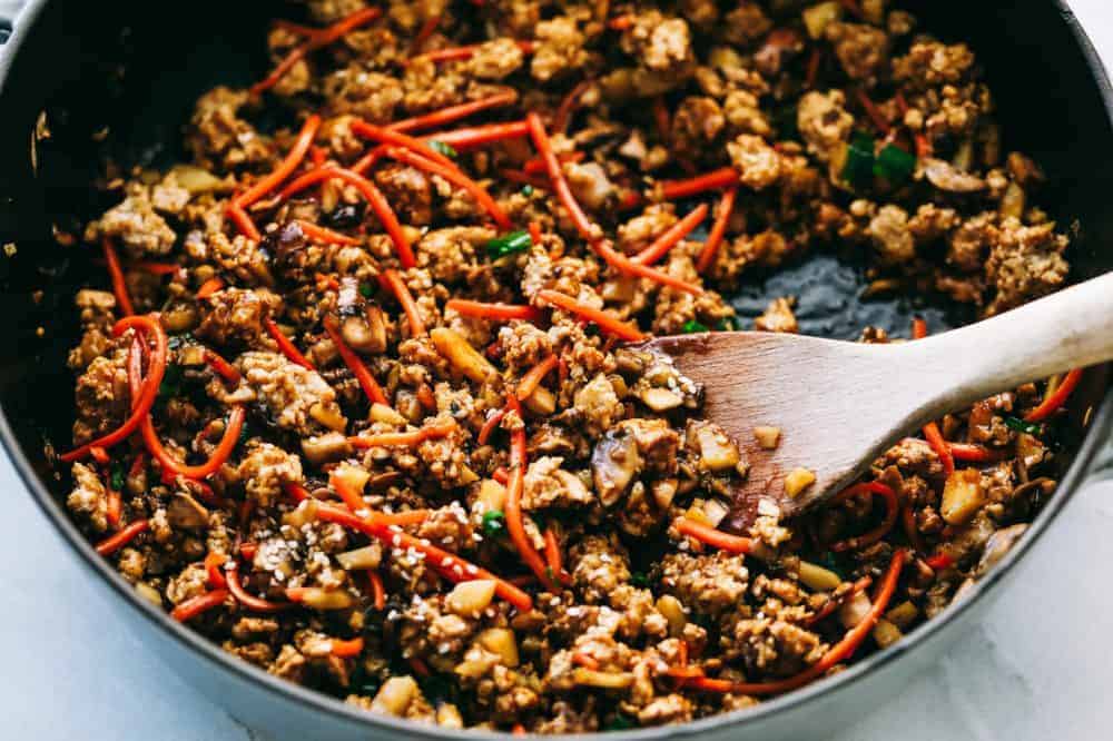 Asian turkey lettuce wrapsย are packed with intense flavorย from tender meat and seasoned, crunchy veggies. These light and tasty wraps are the perfect healthy appetizer or even dinner! The filling for asian turkey lettuce wraps in a skillet with a wooden spoon.