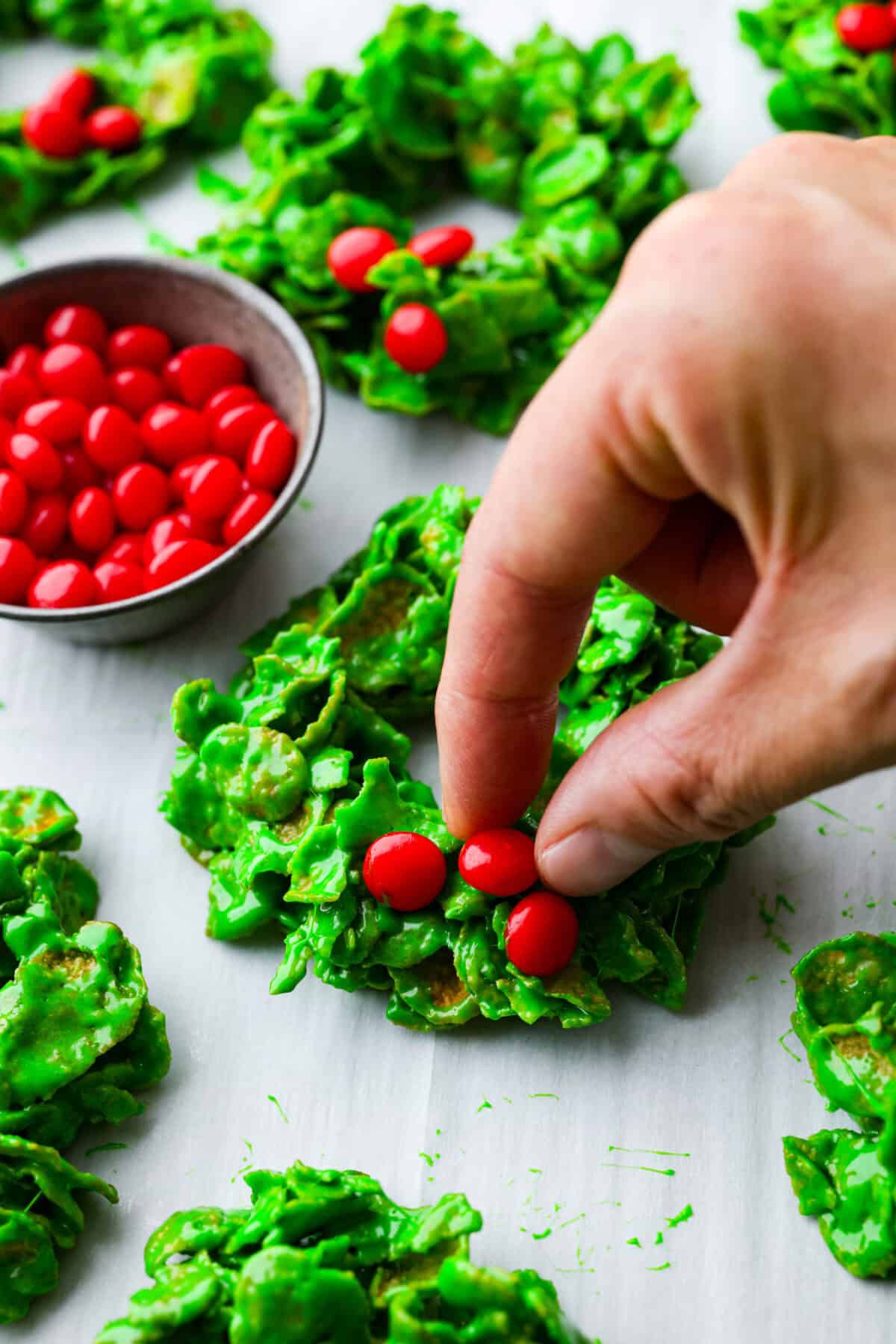 Christmas Cornflake wreaths are a fun, whimsical treat that symbolizes infinite love during the holiday season. Crunchy cornflakes, bright green food coloring, and a sweet, gooey glaze combine to make the cutest Christmas wreath treats. Close up shot of someone placing the cinnamon candies onto the Christmas cornflake wreaths.