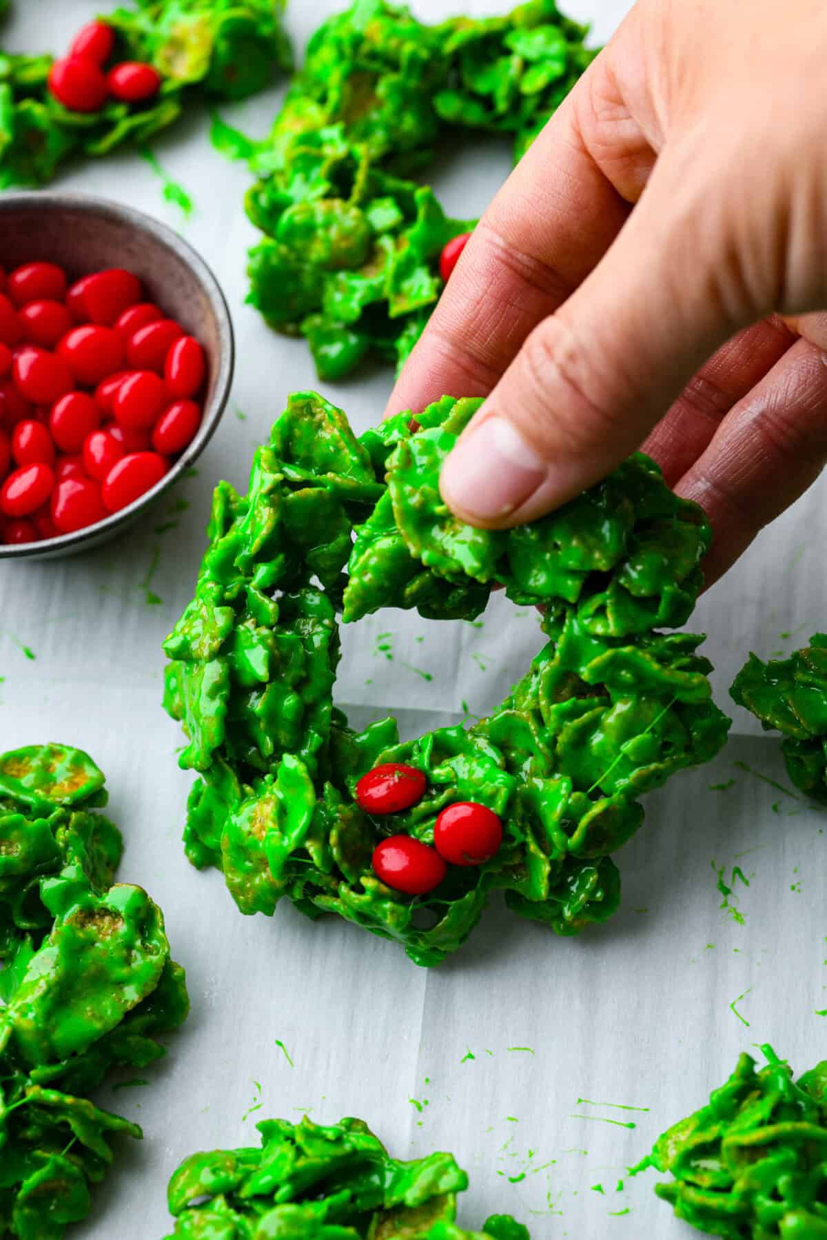 Christmas Cornflake wreaths are a fun, whimsical treat that symbolizes infinite love during the holiday season. Crunchy cornflakes, bright green food coloring, and a sweet, gooey glaze combine to make the cutest Christmas wreath treats. Close up shot of someone picking up a Christmas cornflake wreath.