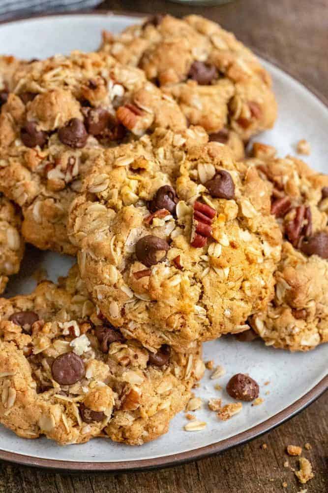 Cowboy Cookies are thick and chewy treats loaded with chocolate chips, oats, shredded coconut, pecans, and cinnamon. Incorporating browned butter into the cookie dough enhances the nutty, caramel notes of the cookie. This recipe packs delightful flavors and textures sure to satisfy any sweet tooth. Cowboy cookies on a white plate.
