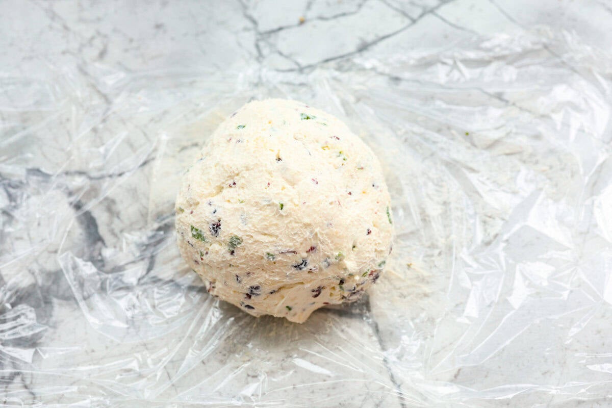 This festive cranberry pecan cheese ball is an easy appetizer for the holiday season! Everyone will love this creamy cheese ball loaded with delicious texture from the dried cranberries and pecans. Serve this impressive appetizer with crackers and enjoy! Fourth photo of the cheeseball formed on a piece of Saran Wrap.