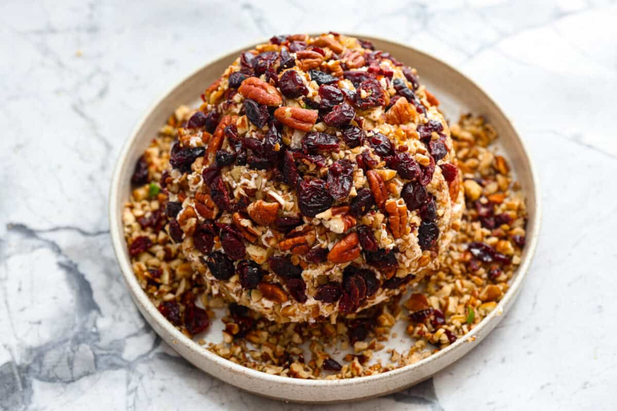 This festive cranberry pecan cheese ball is an easy appetizer for the holiday season! Everyone will love this creamy cheese ball loaded with delicious texture from the dried cranberries and pecans. Serve this impressive appetizer with crackers and enjoy! Sixth photo of the remaining cranberries and pecans being placed on the cheese ball.