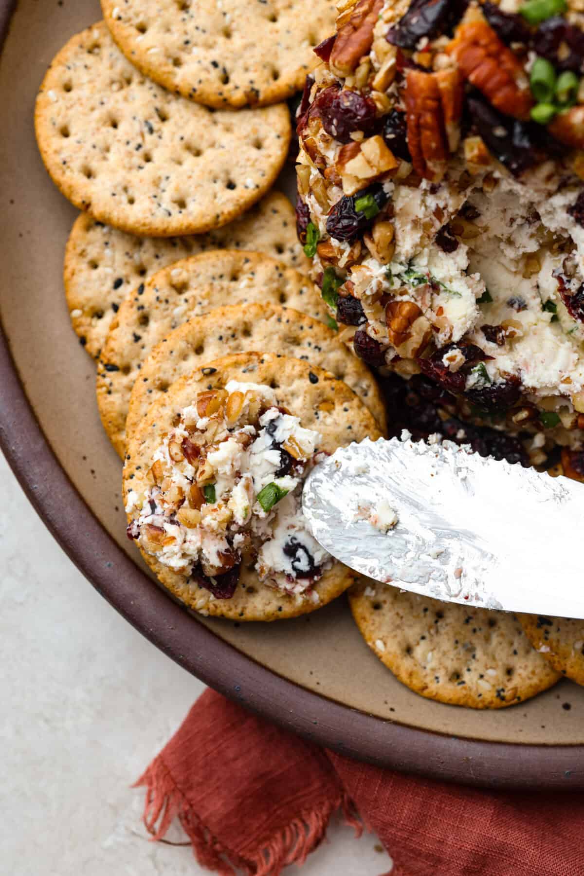 This festive cranberry pecan cheese ball is an easy appetizer for the holiday season! Everyone will love this creamy cheese ball loaded with delicious texture from the dried cranberries and pecans. Serve this impressive appetizer with crackers and enjoy! Top close view of a knife spreading cheese ball on a cracker.