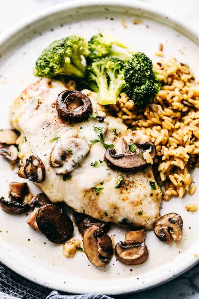Easyย Baked Cheesy Mushroom Chickenย has a melted cheese covering the tender chicken breast with flavorful mushroomsย and mushroom sauce to enhance every bite. The cheese melts and blend the chicken and mushroom flavors together making a meal that isย unforgettable.ย Preparingย this meal is a breeze and givesย you more time to spend with your family!ย Easyย Baked Cheesy Mushroom Chickenย on a plate with rice and broccoli.