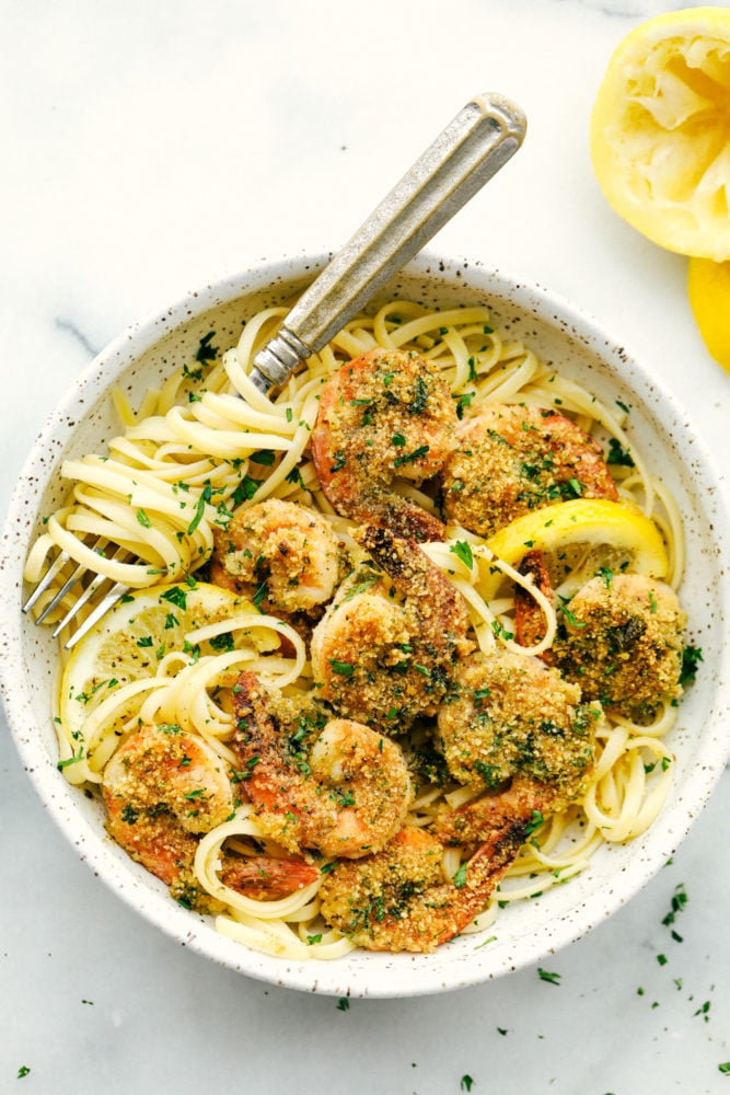 This Lemon Garlic Shrimp Scampi Recipe is a delicious 30 minute meal that cooks in a buttery, lemon garlic sauce and is breaded with a parmesan bread crumb topping. Combined, this meal is irresistible! A true restaurant quality meal right at home! Lemon garlic shrimp in a bowl over top linguine noodles with a fork inside the bowl.