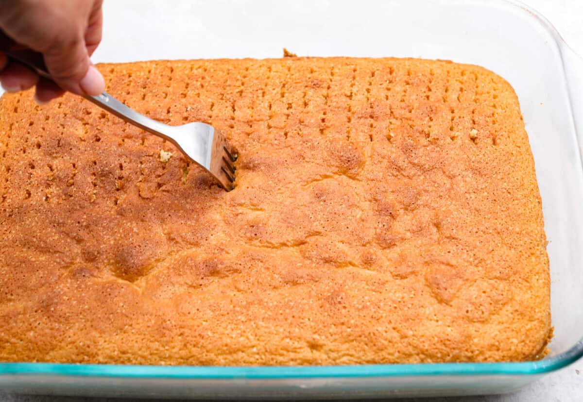 This tender Tres Leches cake is drenched in a three-milk mixture and is topped with fluffy whipped cream, cinnamon, or fresh fruit for a truly authentic Mexican dessert. Angle shot of someone poking the tres leches cake repeatedly with a fork.